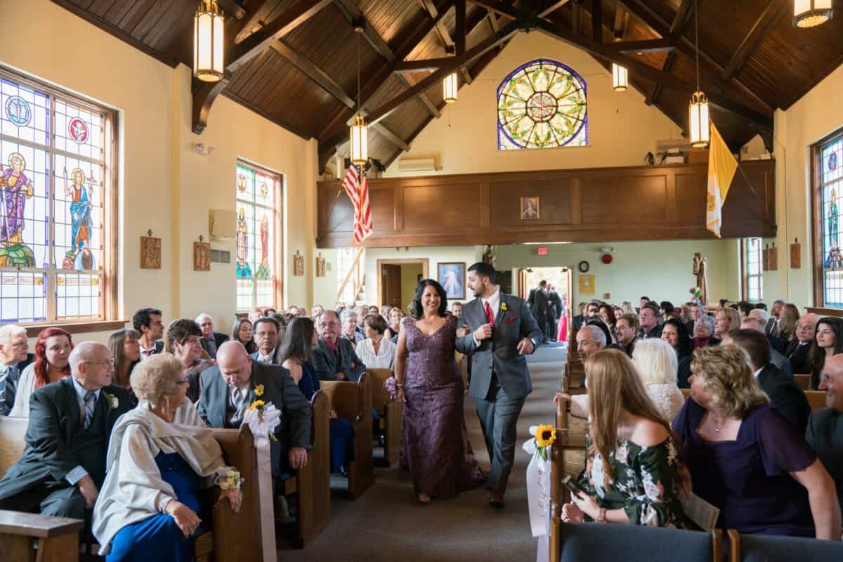 Mother of Bride walks down a church aisle at a Hudson Valley Wedding At West Hills Country Club In Middletown, New York