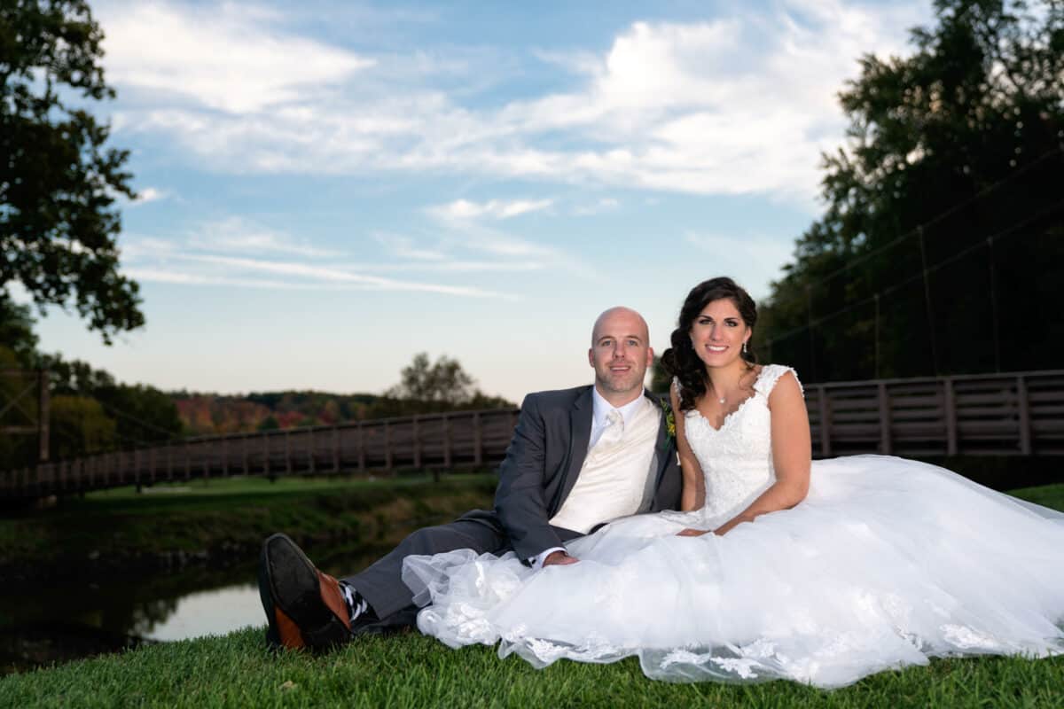 Bride and Groom lay on the grass by a bridge at a Hudson Valley Wedding At West Hills Country Club In Middletown, New York