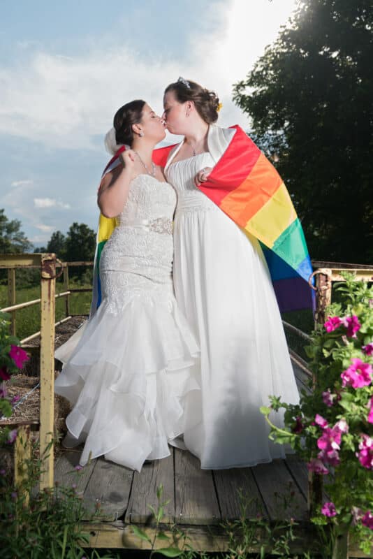 Bride and bride kiss on rustic cart with a rainbow pride flag at Hudson Valley same sex wedding At Lippincott Manor in Walkill New York