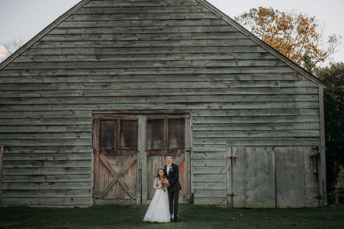 Bride and Groom pose for a photo by a red wooden barn at a wedding at Highlands Country Club